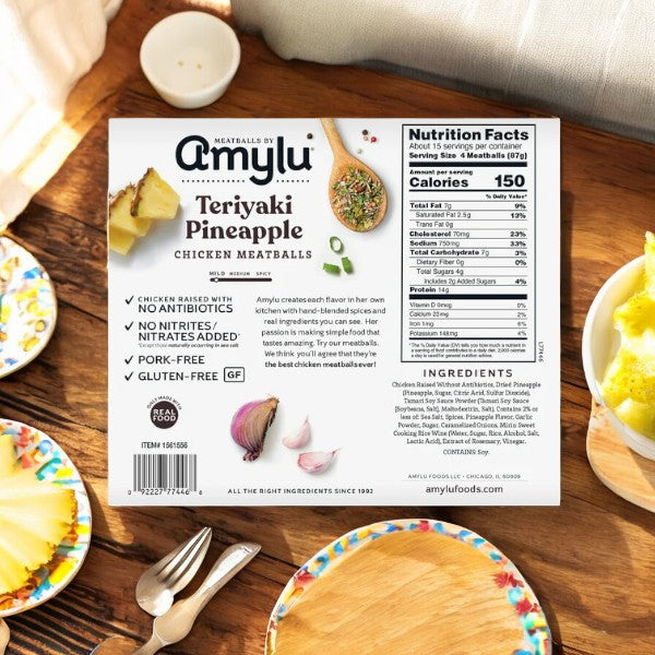 The packaging of Amylu Teriyaki Pineapple Chicken Meatballs, a 46 oz product made with all-natural ingredients, sits on a wooden table surrounded by colorful plates and pineapple slices.