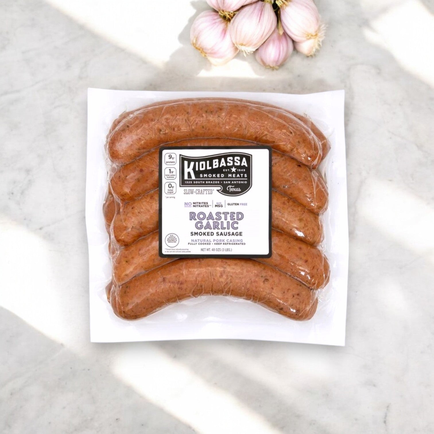 The Kiolbassa Roasted Garlic Smoked Sausage, 48 oz. – Bold, Smoky & Flavor-Packed sits on marble, releasing its slow-smoked aroma with garlic cloves in the background.