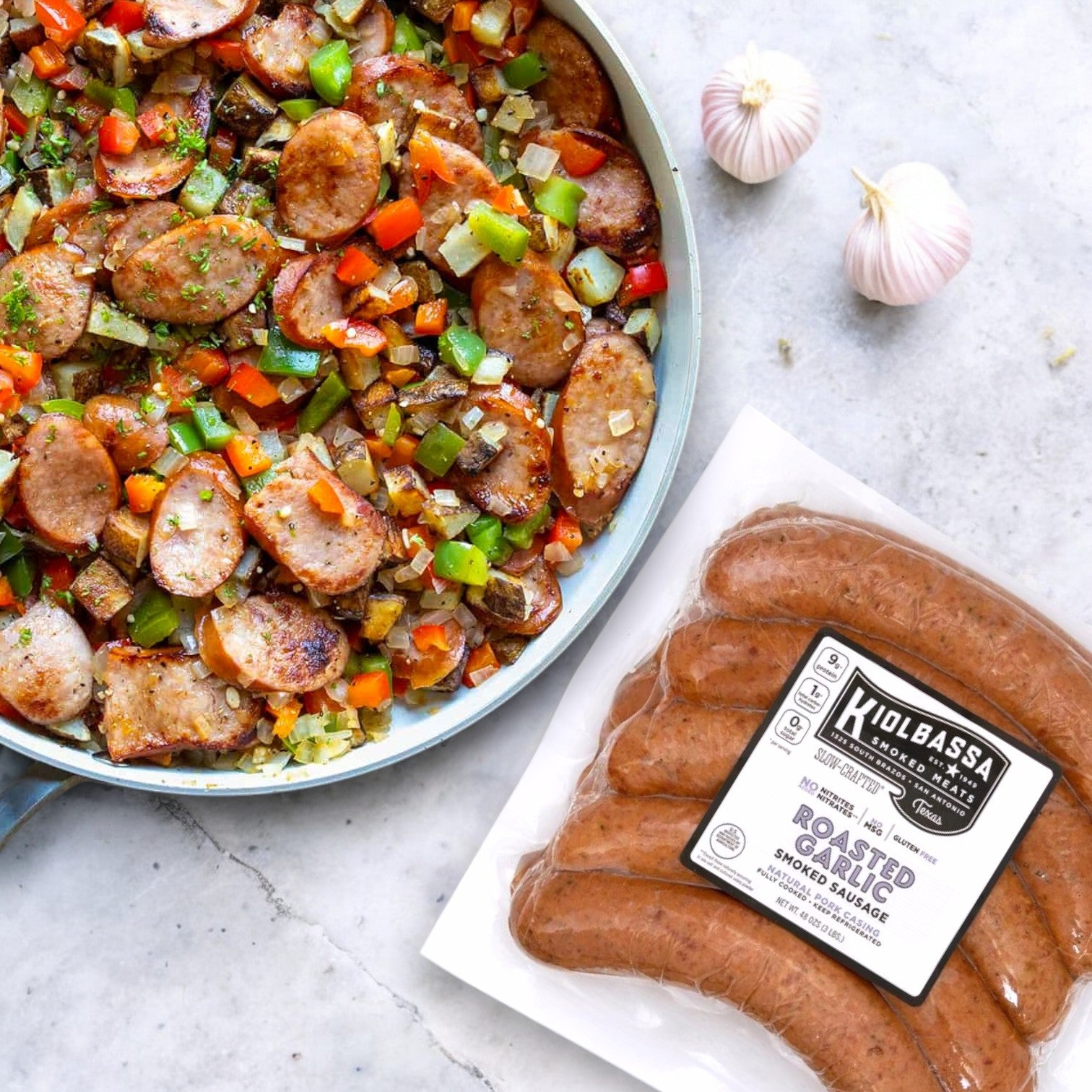 A skillet with sliced sausage, bell peppers, and onions sits next to a 48 oz. package of Kiolbassa Roasted Garlic Smoked Sausage on marble. This premium pork product offers a bold, smoky flavor enhanced by two garlic bulbs nearby.