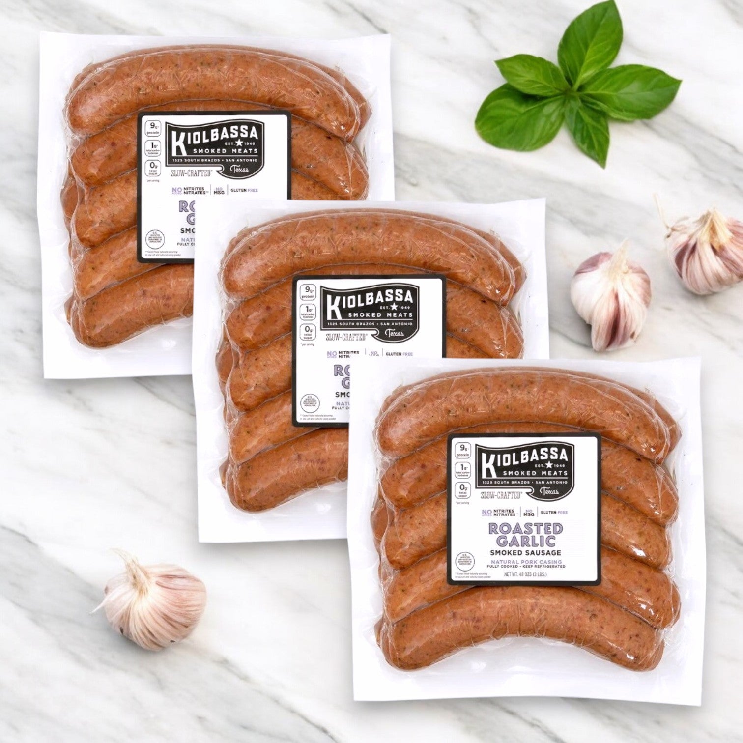 Three vacuum-sealed packs of Kiolbassa Roasted Garlic Smoked Sausage, made with premium pork and offering a bold, smoky flavor, sit on a marble surface surrounded by garlic cloves and basil.