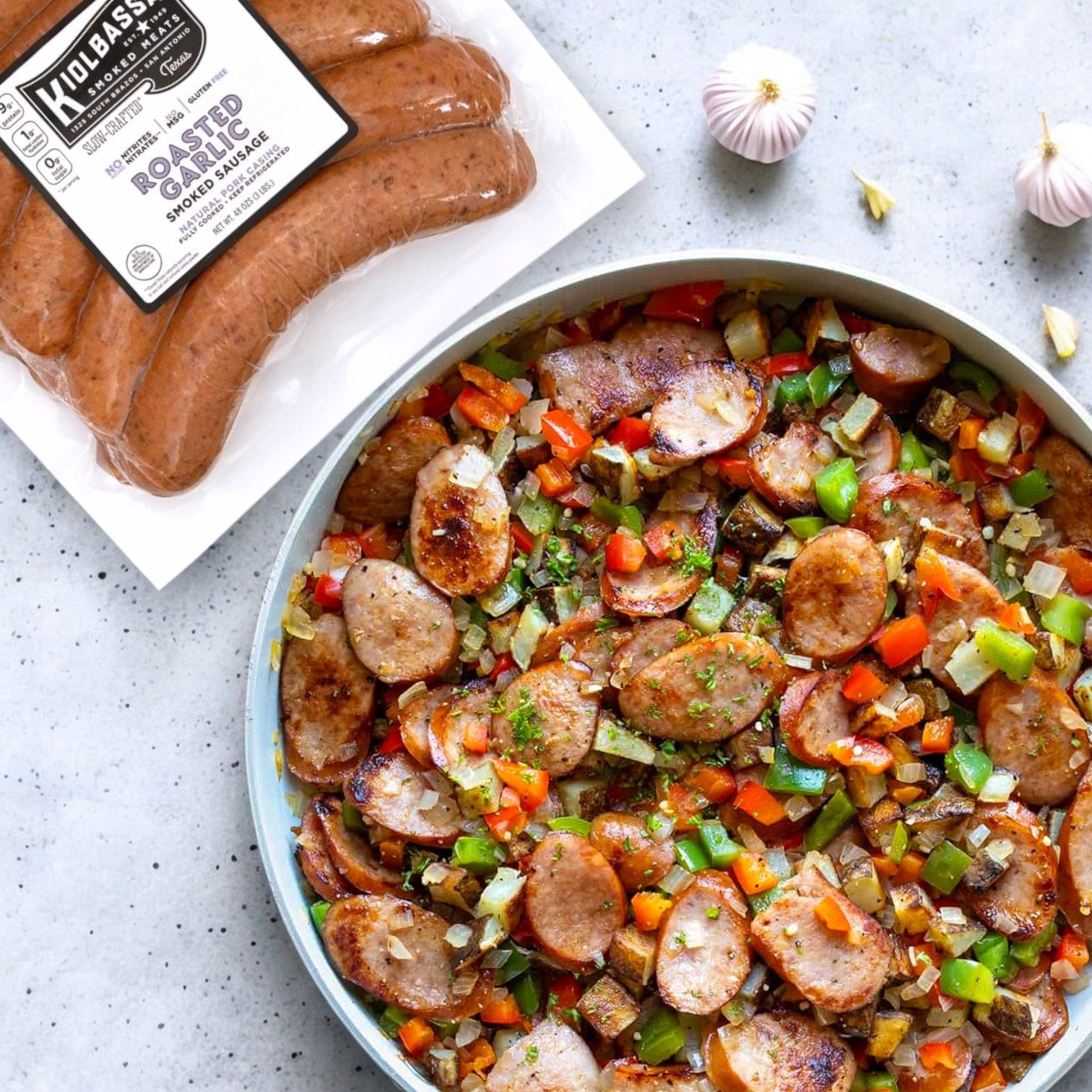 On a gray surface, skillet-cooked slices of Kiolbassa Roasted Garlic Smoked Sausage, 144 oz.—bold, smoky & flavorful—are mixed with diced peppers and onions, nestled next to premium pork sausages and garlic cloves.