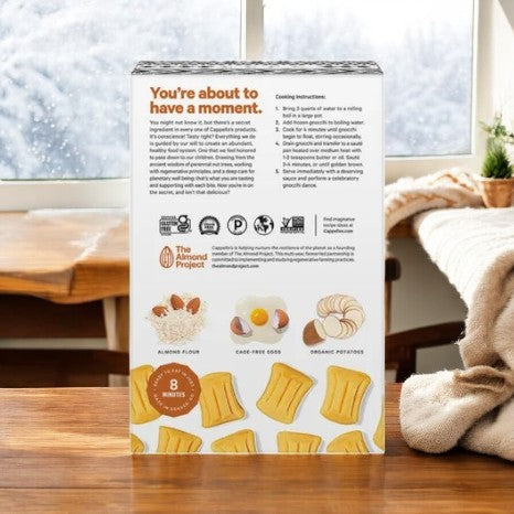 On a wooden table, the back of the Cappello's Gnocchi Pasta package displays cooking instructions along with images of gluten-free ingredients such as almond flour, cage-free eggs, and organic potatoes. The text reads "You're about to have a moment. Perfectly paleo-friendly.