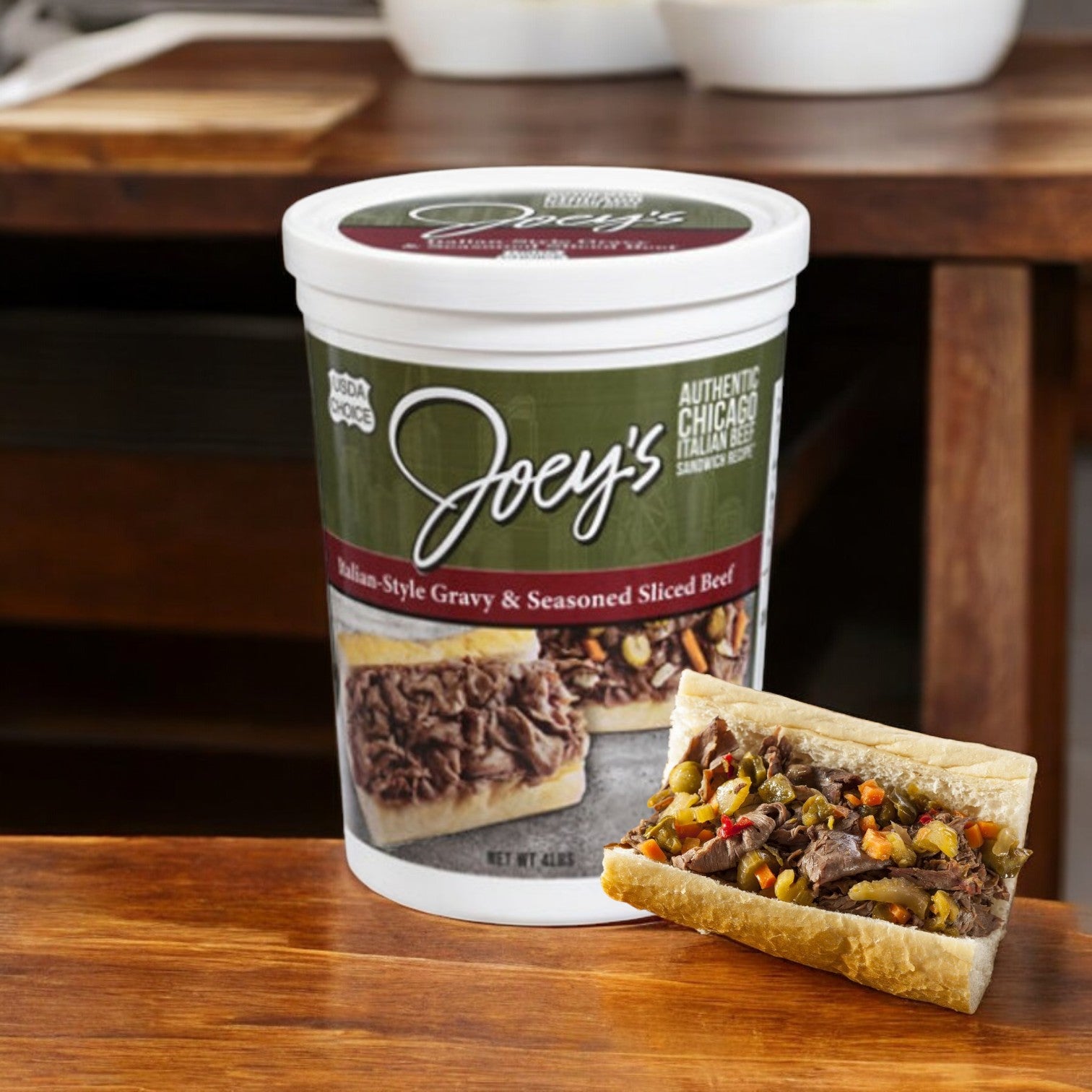 A "Joey's Sliced Beef, Gravy & Seasoned, Italian-Style" tub from Easy Lunches rests on a wooden table next to a sandwich filled with seasoned sliced beef and fresh vegetables.