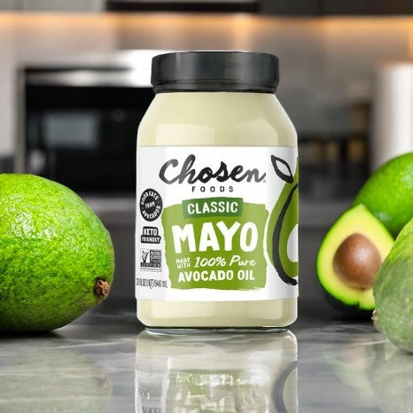 A 32 fl oz jar of Chosen Foods Avocado Oil Classic Mayo, made with pure avocado oil, rests on a kitchen countertop amid whole and halved avocados. This gluten-free mayonnaise adds rich flavor, ideal for enhancing your favorite dishes.