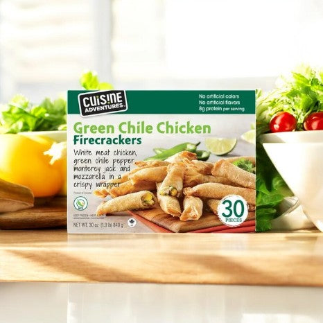 A box of 30 Cuisine Adventures Green Chile Chicken Firecrackers, each encased in a crispy golden shell, is displayed alongside fresh vegetables on a wooden surface. Ideal for anyone seeking a boldly flavorful snack.