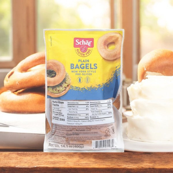 A bag of Schär New York Style gluten-free plain bagels sits on a wooden surface, capturing the essence of NYC. In the background, bagels and cream cheese beckon those with gluten sensitivities to enjoy without compromise.