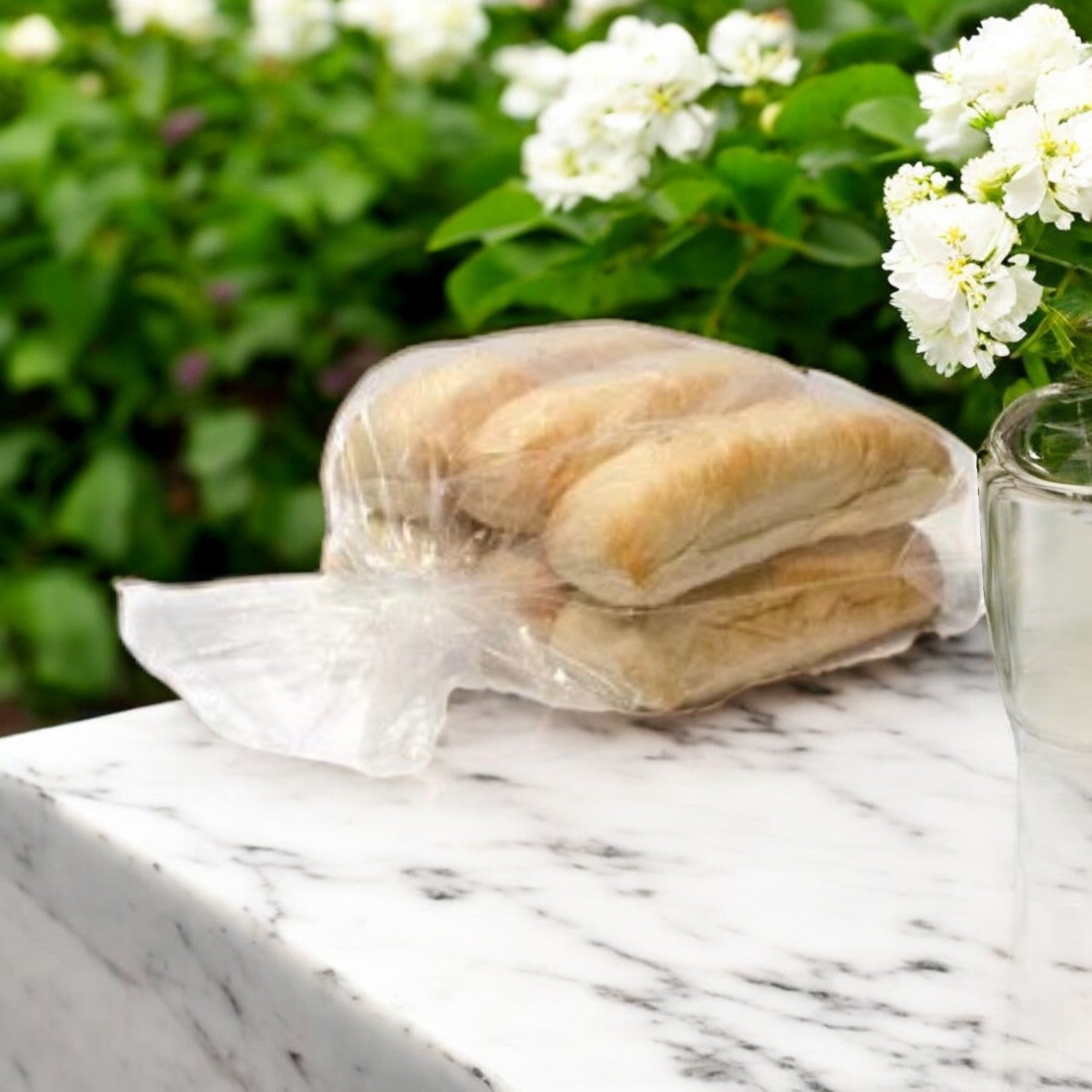 A plastic bag with baguettes is on a marble surface beside a vase of white flowers, perfect for crafting an Italian beef sandwich using the Buona Marinara Sauce & Meatballs Kit + Italian Beef -12 Total Sandwiches! Greenery in the background hints at freshness.