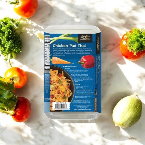 Snapdragon Chicken Pad Thai (32 oz, 1 Count) sits on a marble surface amidst fresh broccoli, tomatoes, and peppers. This frozen meal by Snapdragon brings an authentic Pad Thai experience straight to your kitchen.