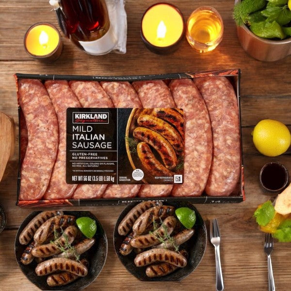 A pack of Kirkland Signature Mild Italian Sausage-1 Count rests on a wooden table, accompanied by two plates featuring cooked sausages, candles, lemons, seasonings, and a salad, creating a cozy meal setting.