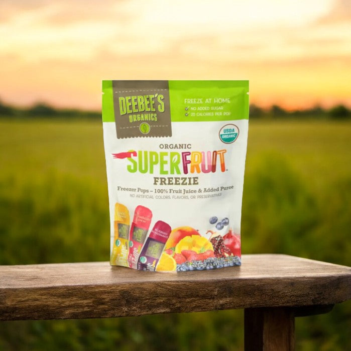 A bag of Deebee's DeeBee's Organics Freezie Pops, Organic - 10 -1.35 fl oz rests on a wooden surface, set against a blurred background of a grassy field. The packaging highlights its organic, plant-based ingredients and absence of added sugar.