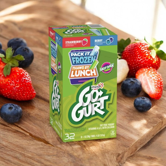 A box of Yoplait Go-Gurt Simple Low Fat Yogurt, Variety, 2.25 oz, 32 Count, in strawberry and berry flavors, is placed on a wooden surface alongside fresh strawberries and blueberries. The packaging highlights "Pack it frozen, thaws by lunch." Perfect for a kids snack on-the-go!