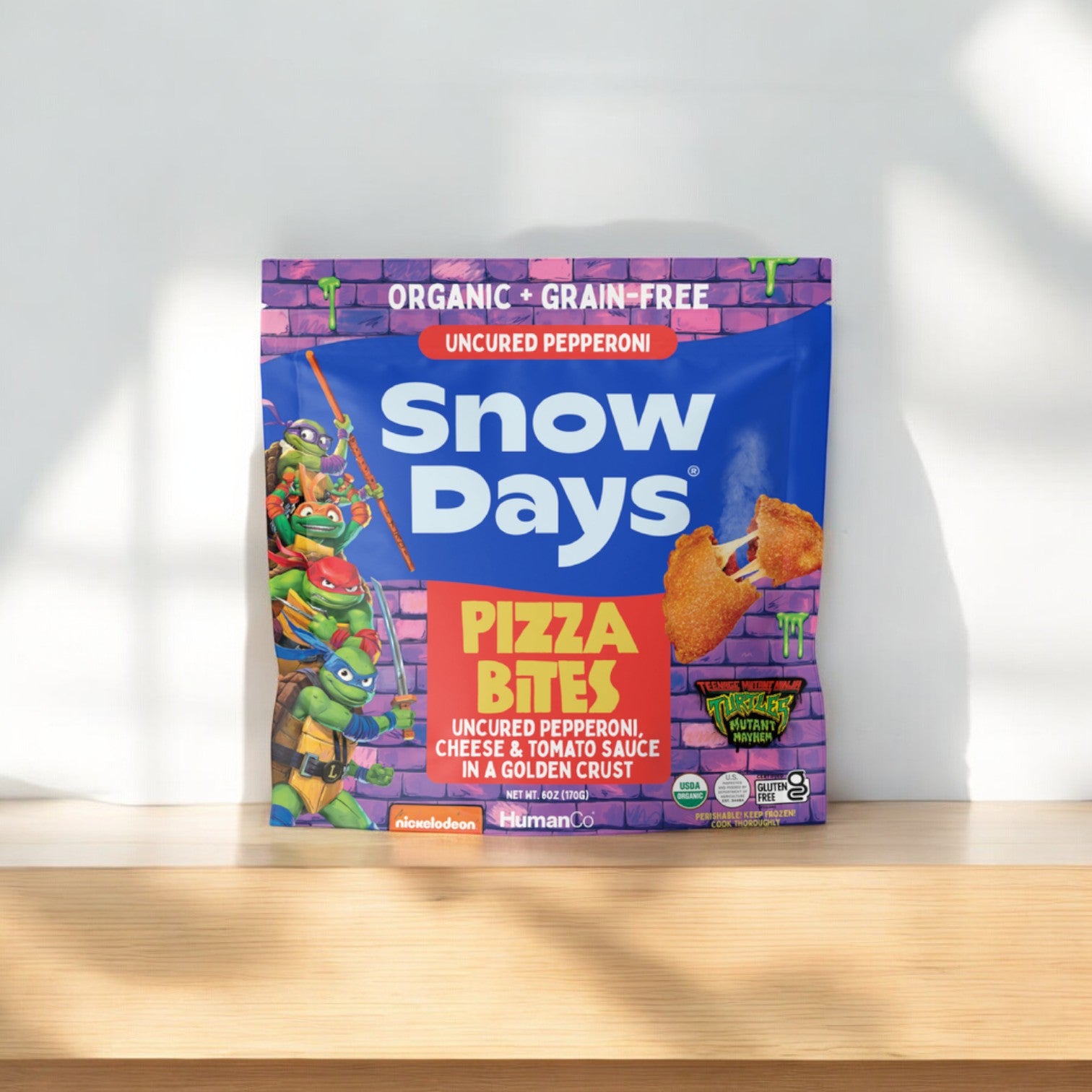 Snow Days Uncured Pepperoni Pizza Bites come in a 6 oz. bag and are gluten-free and grain-free, featuring cheese and tomato sauce in a golden crust. The packaging showcases cartoon characters and emphasizes organic ingredients that are high in protein.