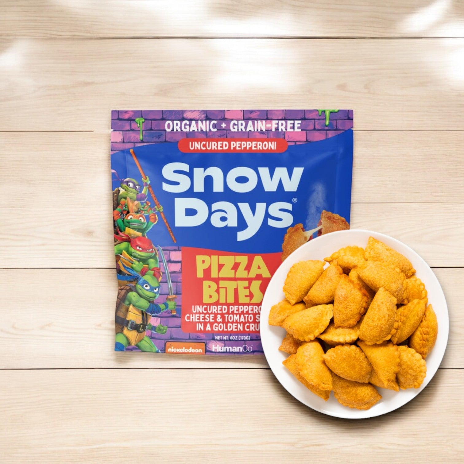 A bag of Snow Days Uncured Pepperoni Pizza Bites, which are gluten and grain-free, accompanies a plate full of these tasty treats. The packaging showcases cartoon characters, and the bites offer a protein-rich option for a satisfying snack.