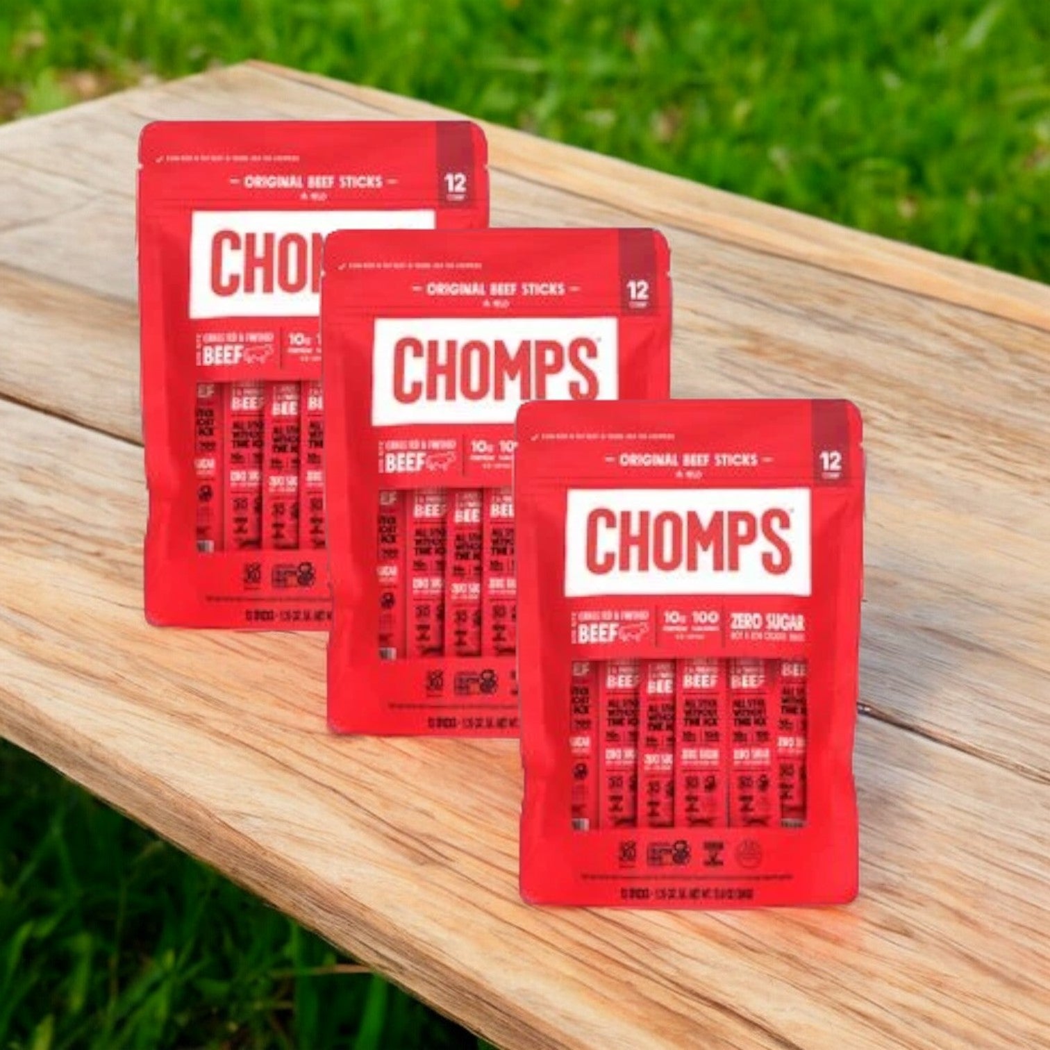 Three bags of Chomps Original Beef, totaling 36 gluten-free meat sticks, are arranged on a wooden surface with a grassy background.