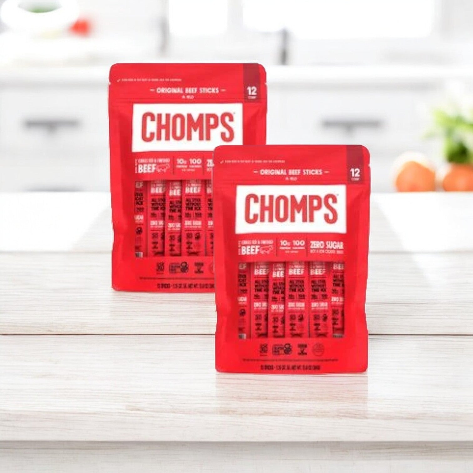 Two packages of Chomps Original Beef 12 Pack, each containing 12 high-protein, sugar-free beef sticks made from grass-fed beef, are displayed on a wooden surface with a kitchen background.