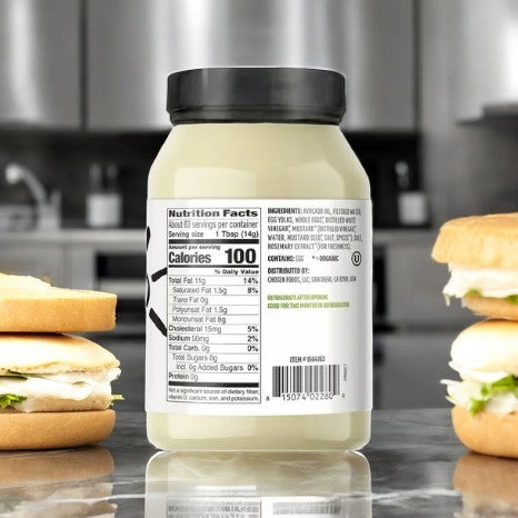 A jar of Chosen Foods Avocado Oil Classic Mayo (32 fl oz) is on the counter, surrounded by two sandwiches. Its label highlights nutrition facts and ingredients, showcasing its healthy mayonnaise components.