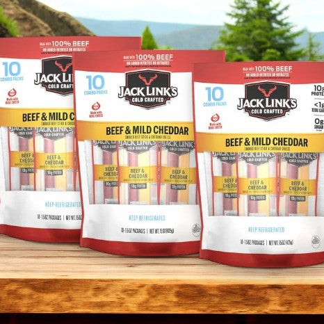 The 3-count packs of Jack Link's Cold Crafted Beef Stick & Mild Cheddar Cheese Combo are arranged on a wooden surface, with a scenic backdrop of trees and mountains, ideal for enjoying a quick snack while immersed in nature.