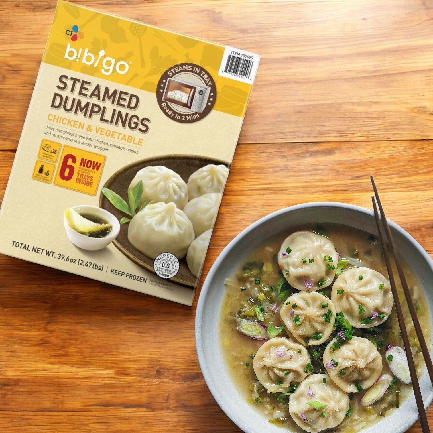 A box of CJ Bibigo Steamed Dumplings, Chicken & Vegetable (39.6 oz, 6-count), is beside a bowl of steaming chicken and vegetable dumplings in broth, garnished with herbs, on a wooden surface.