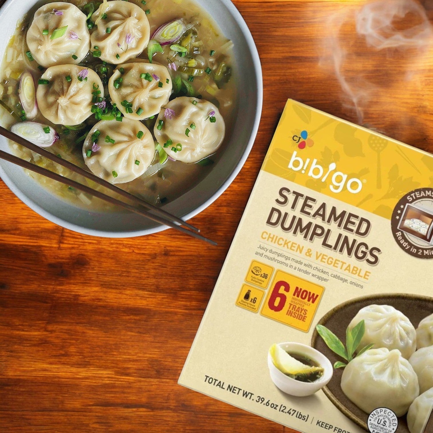 A bowl of Korean-style dumplings in broth with scallions is next to a box of CJ Bibigo Steamed Dumplings, Chicken & Vegetable, 39.6 oz (18-count), on a wooden surface.