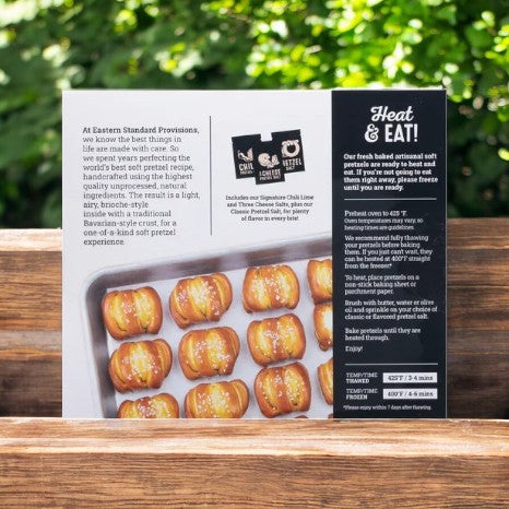 A box of Eastern Standard Provisions Pretzel Bites- 1 Count, crafted with high-quality ingredients, rests on a wooden surface amid lush greenery. Heating instructions on the back promise a delightful, chewy snack experience.