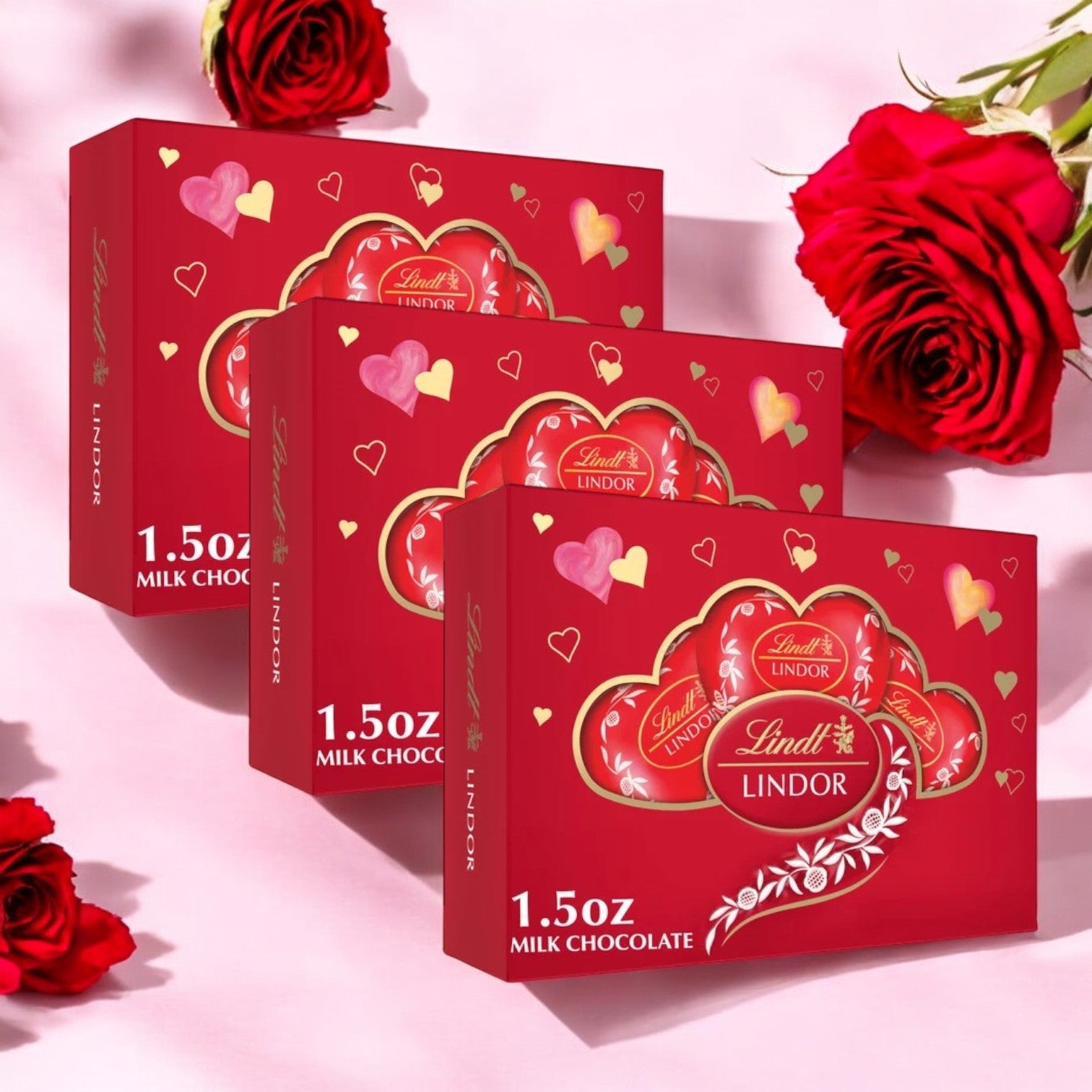 Displayed against a background of red roses are three Lindt Lindor Valentine's Milk Chocolate Truffles Heart Gift Boxes, each weighing 1.5 oz.