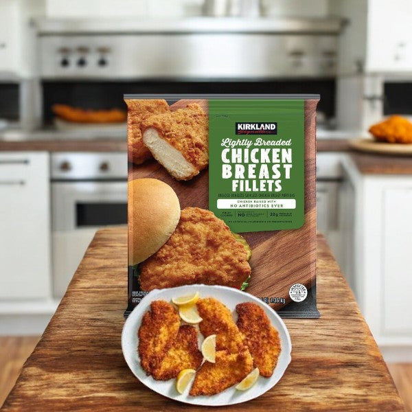 A Kirkland Signature Lightly Breaded Chicken Breast Fillet from a 3-lb box rests on the kitchen counter, while a plate of lean, cooked fillets garnished with lemon slices is displayed in the foreground.