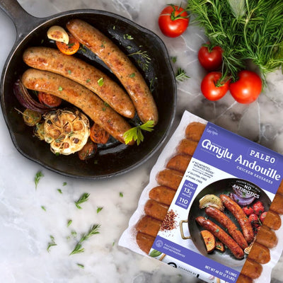 A skillet of cooked sausage, roasted garlic, and tomatoes sits beside a pack of Amylu Paleo Andouille Sausage, 16 Count. The Cajun-inspired flavor pops as fresh dill and juicy tomatoes elegantly adorn the marble surface for a tasty paleo-friendly dish.