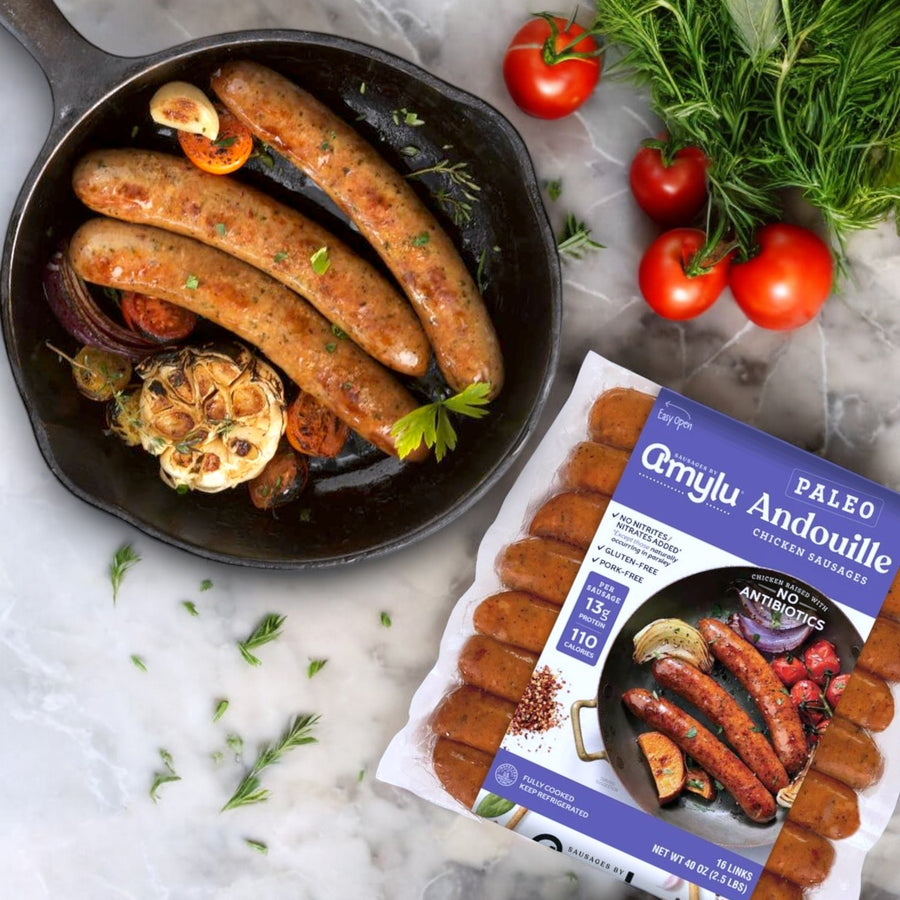 A skillet of cooked sausage, roasted garlic, and tomatoes sits beside a pack of Amylu Paleo Andouille Sausage, 16 Count. The Cajun-inspired flavor pops as fresh dill and juicy tomatoes elegantly adorn the marble surface for a tasty paleo-friendly dish.