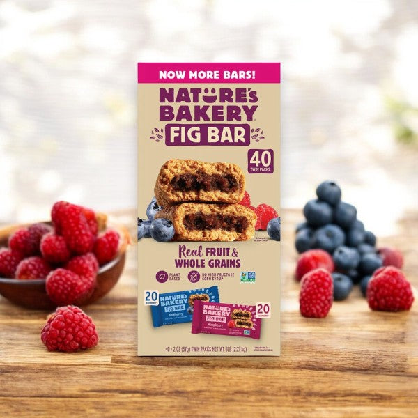 A box of Nature's Bakery Fig Bar, Variety Pack, 2 oz, 40 Count by Easy Lunches is displayed on a wooden table with fresh raspberries and blueberries blurred in the background. The box highlights the inclusion of various flavors and real fruit in each of the 40 twin packs.