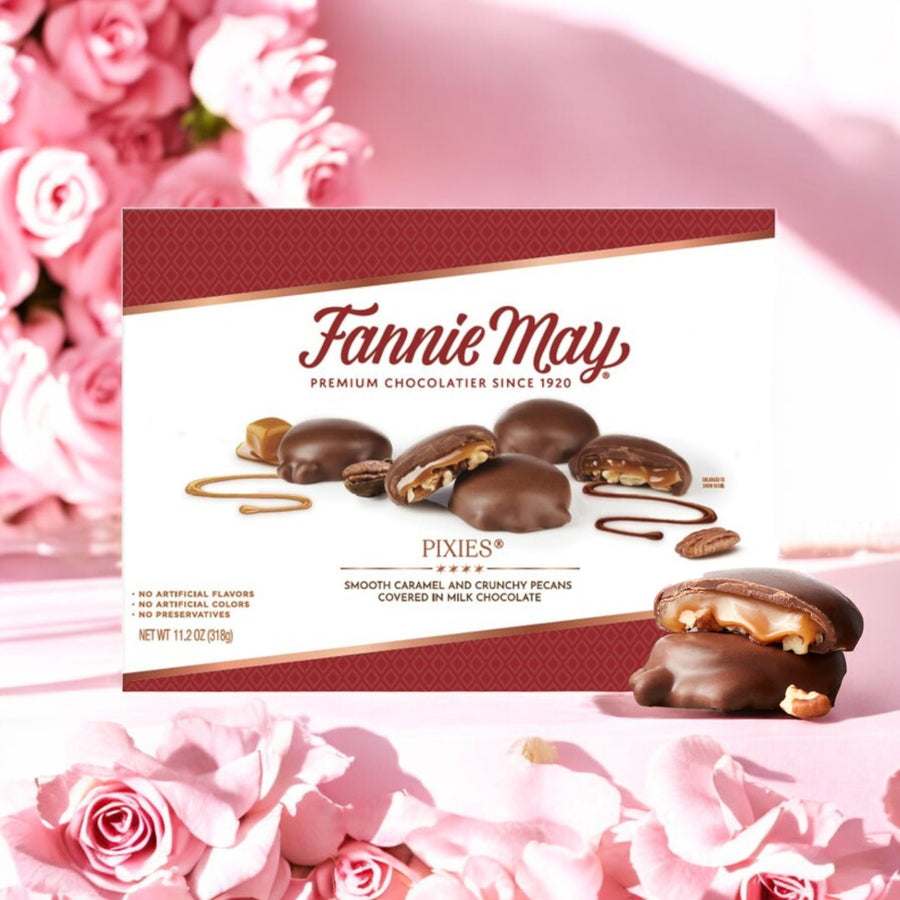 A box of Fannie May Pixies Chocolate – 1 Count- 11.2 oz by Fannie May is shown on a pink surface with pink roses in the background.