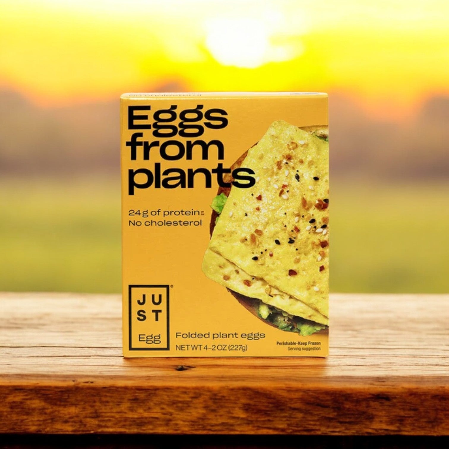 A pack of JUST Egg Folded plant-based eggs by Just Egg sits atop a wooden surface, set against a softly blurred sunset backdrop. The text emphasizes its protein content and highlights this cholesterol-free, plant-based alternative.