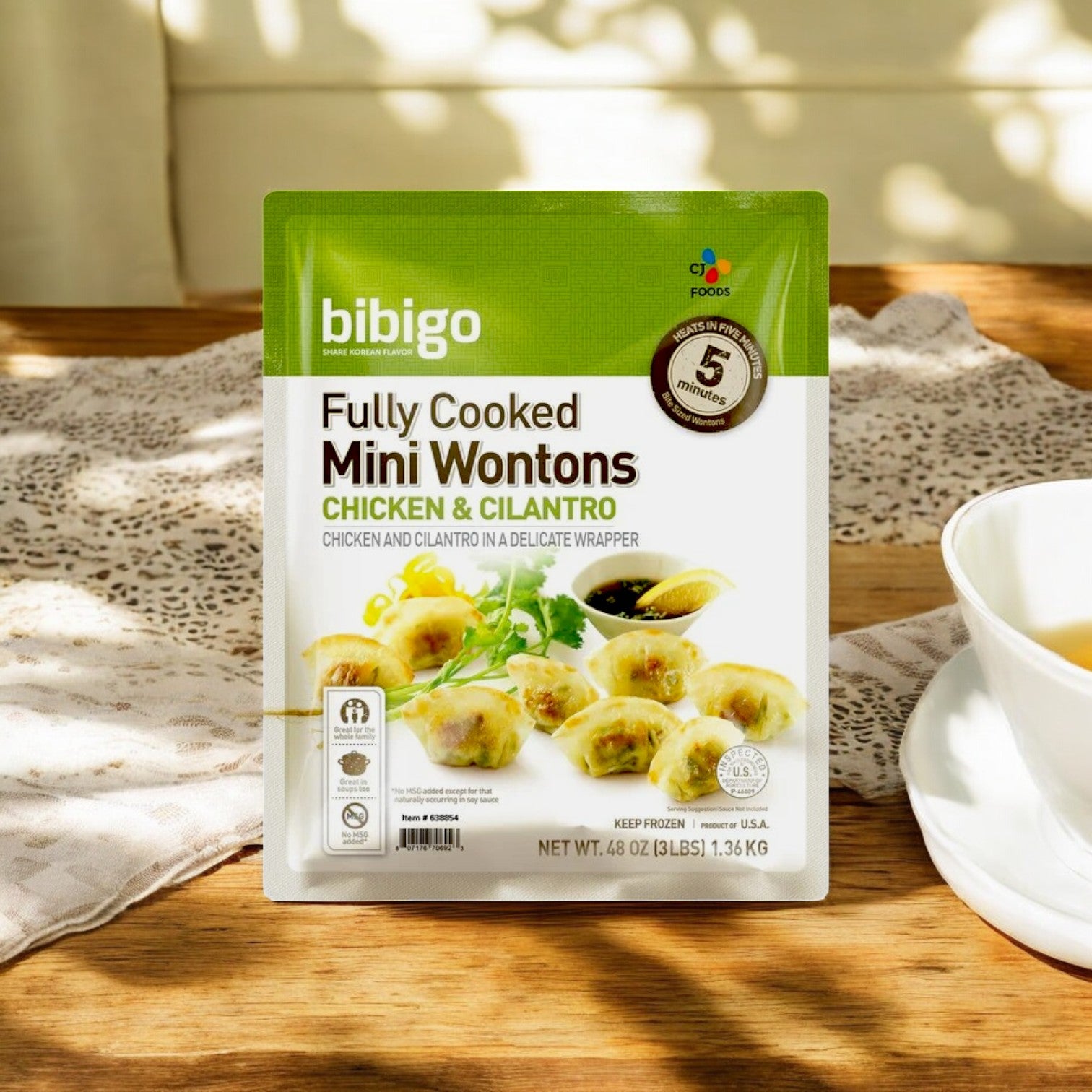 A pack of Bibigo Mini Wontons – Chicken & Cilantro (3 lbs) by CJ Bibigo rests elegantly on a wooden table with a lace doily and cup, providing an authentic Korean taste akin to traditional dumplings.