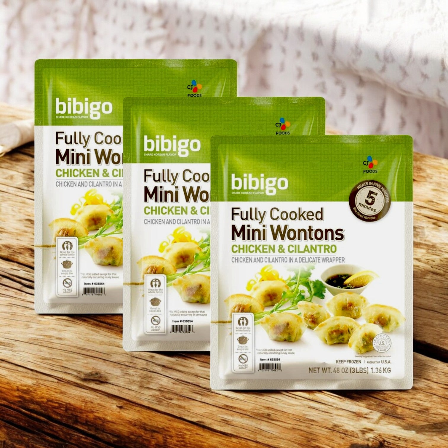 Three packages of CJ Bibigo Mini Wontons – Chicken & Cilantro (3 lbs) are showcased against a wooden background, offering a delightful taste of Korean cuisine.