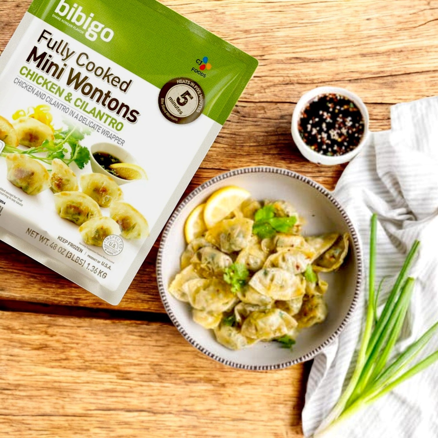 A package of Bibigo Mini Wontons – Chicken & Cilantro (3 lbs) sits beside a plate of wontons with greens and lemon slices. Discover Korean flavors with scallions, bulgogi seasoning, and vibrant spices from CJ Bibigo on a wooden surface.