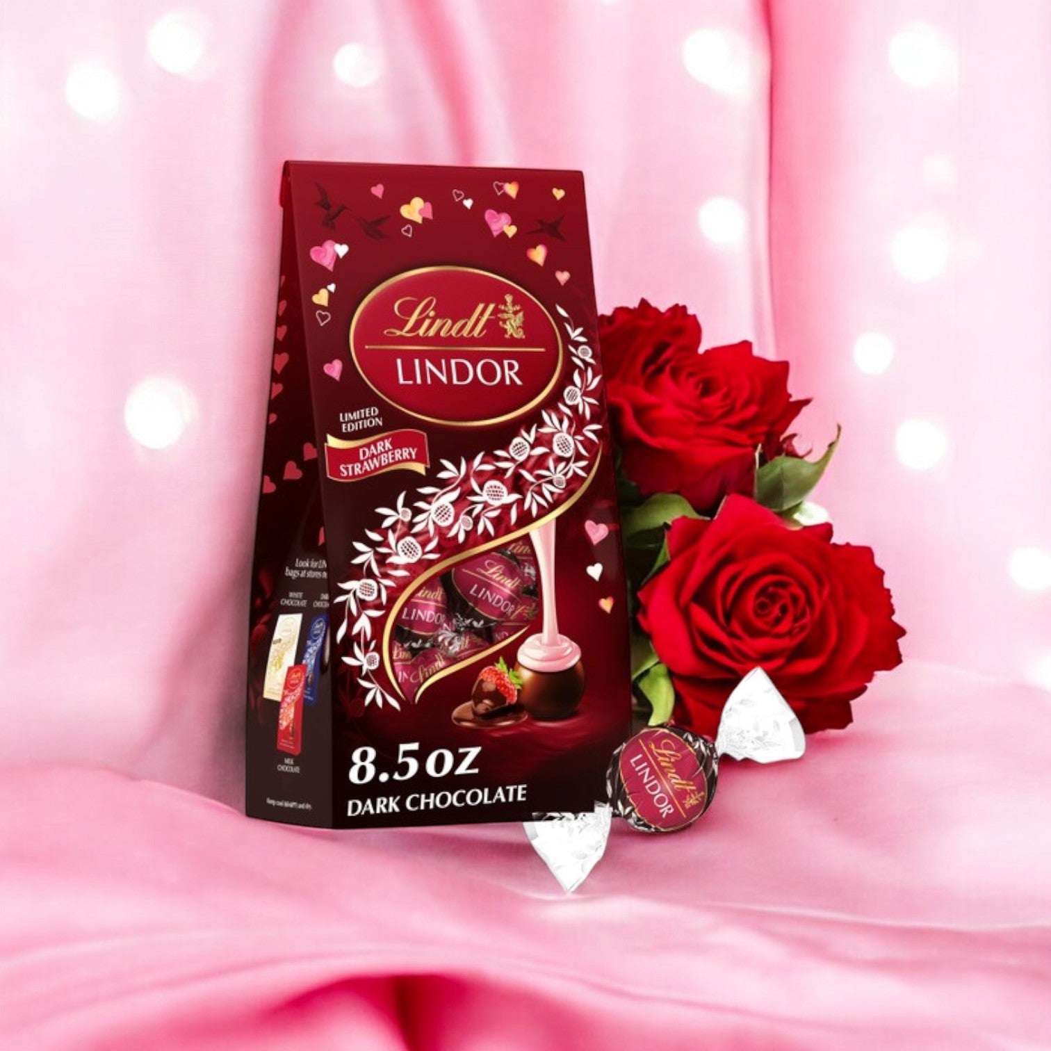 A bag of Lindt Lindor Valentine's Strawberry Dark Chocolate Candy Truffles – 8.5 oz rests on pink fabric amid red roses, creating a perfect Valentine’s Day candy delight.