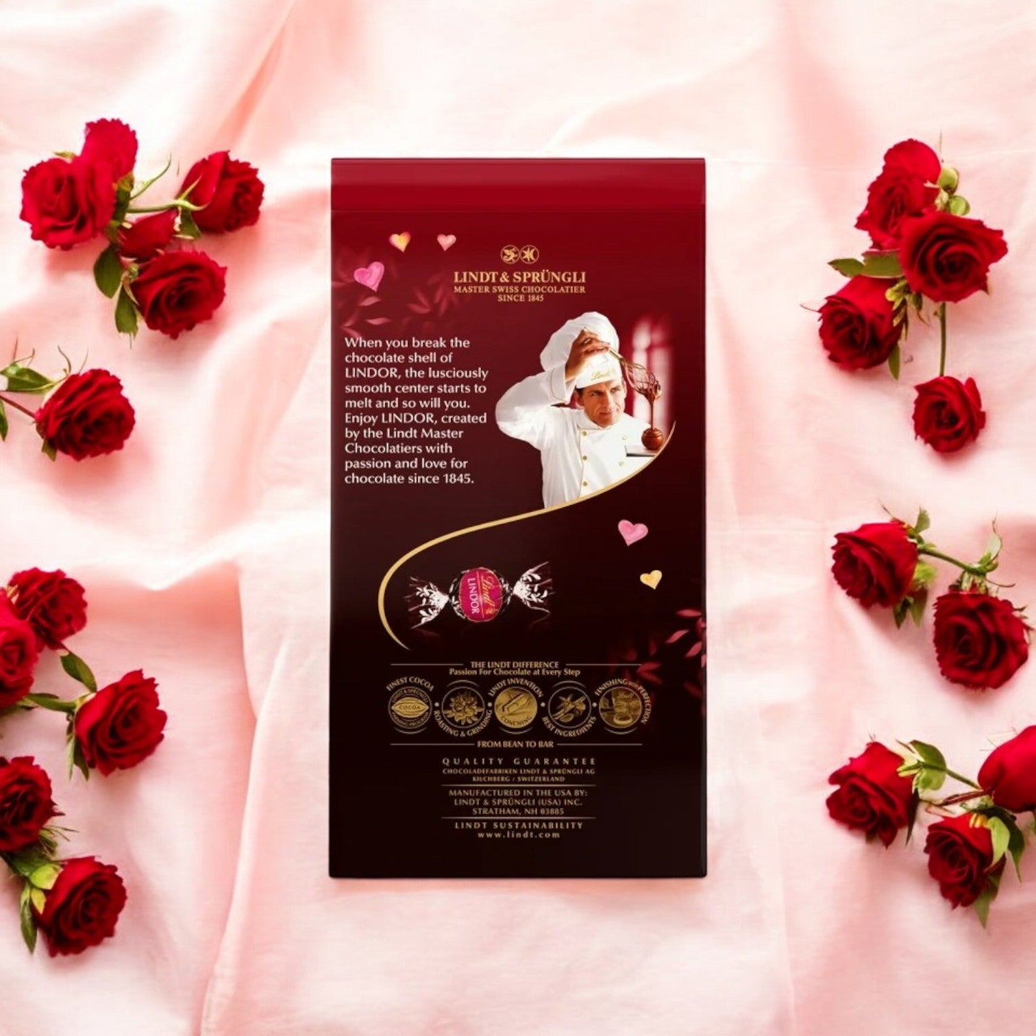 An 8.5 oz package of Lindt Lindor Valentine's Strawberry Dark Chocolate Truffles rests on pink fabric surrounded by red roses, featuring exquisite text and a chocolatier image, promising a decadent experience with dark chocolate truffles.