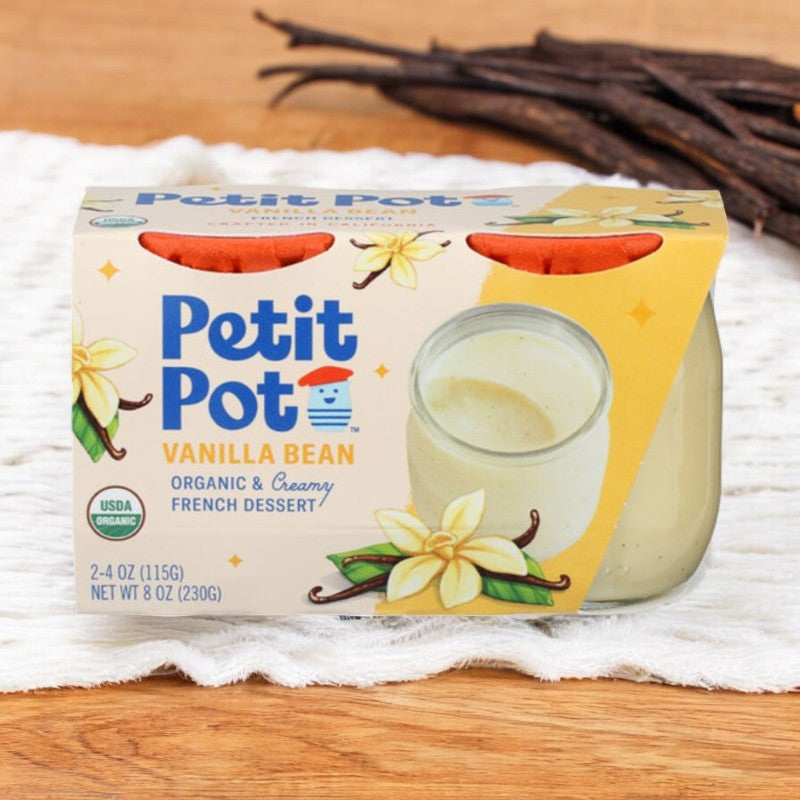 A package of Petit Pot's Organic French Vanilla Pudding - 2 Petit Pots rests on a wooden surface with vanilla beans in the background. The front of the package showcases the organic certification and a dessert image, emphasizing the French Vanilla flavor.