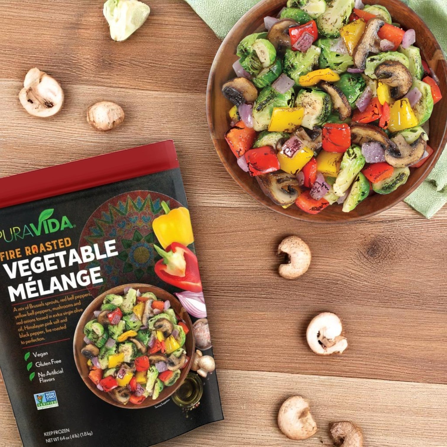A bowl of fire-roasted vegetables sits beside a Pura Vidaa Fire-Roasted Vegetable Mélange – 64 oz Family Pack, displayed on a wooden surface with scattered broccoli and mushroom pieces nearby.