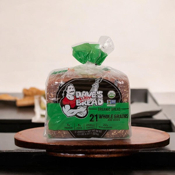 A loaf of Dave's Killer Bread Organic 21 Whole Grain in plastic packaging rests on a wooden surface. The green packaging displays an illustration of a muscular man with a guitar and prominently highlights its USDA Organic and Non-GMO certifications.