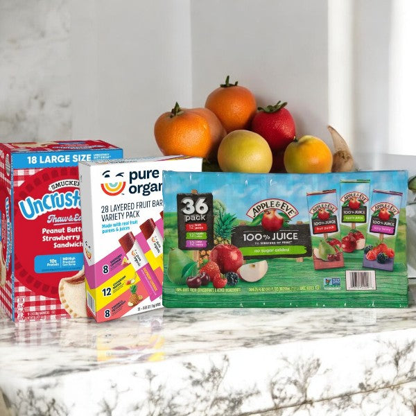 A marble countertop holds a bowl of fruit alongside packs from Easy Lunches' Kid's Bundle, which includes 18 Strawberry Jam Uncrustables, 28 Pure Organic Assorted Fruit Bars, and 36 Apple & Eve Assorted Juice Boxes.