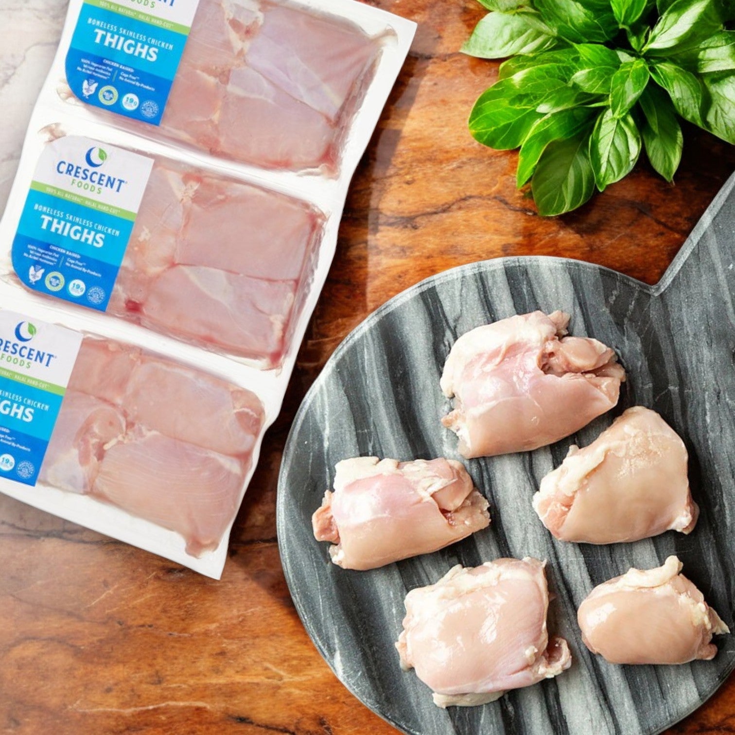 Crescent Foods Halal Chicken Thighs, hand-cut, boneless, and skinless (approx. 4 lbs), both packaged and unpackaged, are placed on a marble surface with fresh basil nearby.