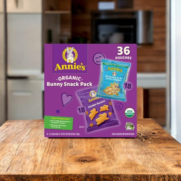 A box of Easy Lunches' Annie's Organic Bunny Snack Pack Baked Crackers and Graham Snacks, containing 36 individually wrapped pouches, sits on a wooden table in a kitchen setting. The packaging showcases a variety pack with two types of snacks: 18 Birthday Cake and 18 Cheddar Bunnies, all made with organic ingredients.