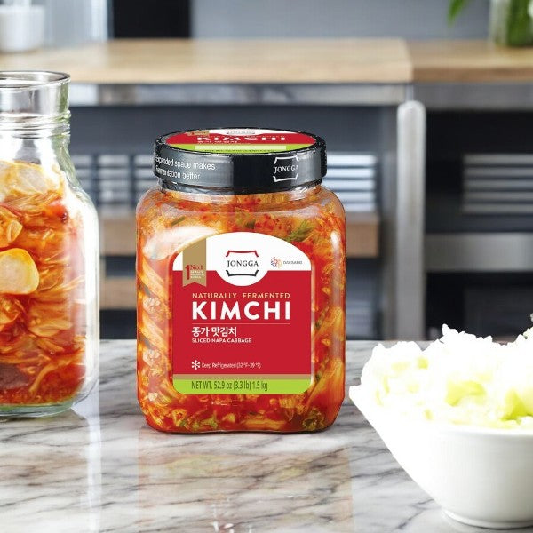 A jar of Kimchi- Jongga Sliced Napa Cabbage Kimchi, 52.9 oz by Easy Lunches rests on a kitchen counter beside a bowl of chopped Napa cabbage.