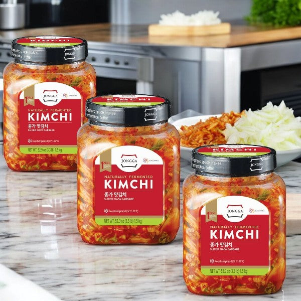 Three jars of Kimchi Jongga Sliced Napa Cabbage Kimchi, 52.9 oz- 3 Pack by Easy Lunches are arranged on a kitchen counter, their vibrant colors accentuated against a backdrop of chopped Napa cabbage kimchi.