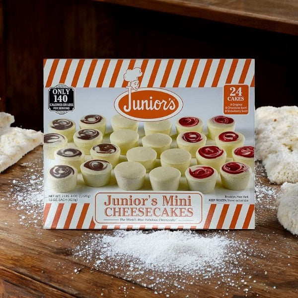 A box of Juniors Mini Cheesecakes, Variety Pack, rests on a wooden surface, displaying an array of New York-style cheesecake indulgences in both plain and strawberry swirl flavors.