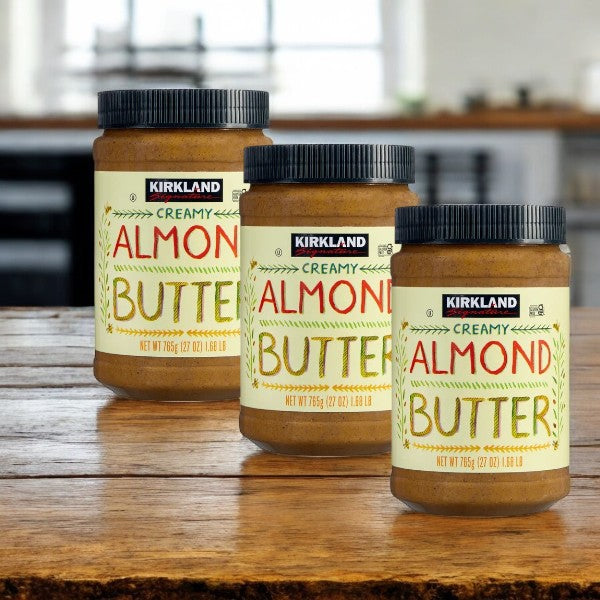 Three jars of Kirkland Signature Creamy Almond Butter (27 oz-3 Count) are arranged on a wooden table, highlighting their gluten-free and high-protein features against a kitchen backdrop.