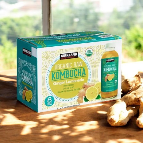 A package of Kirkland Signature Kombucha Organic Ginger Lemonade rests on the table, highlighting a single bottle of probiotic goodness. Fresh ginger in the foreground suggests its invigorating flavor.