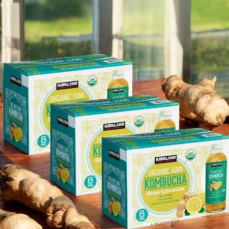 Boxes of Kirkland Signature Kombucha Organic Ginger Lemonade, a 3-count pack celebrated for its probiotic benefits and gut health support, are displayed next to ginger roots in a sunlit room.