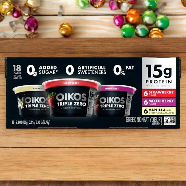 A package of Dannon's Oikos Triple Zero Greek Nonfat Yogurt, presented in an 18-count variety pack featuring vanilla, strawberry, and mixed berry flavors, is set against a wooden background. This non-GMO delight offers 15g of protein, contains no added sugars, and serves as a good source of calcium.