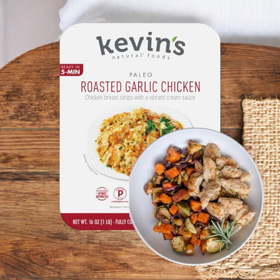 Packaging of Easy Lunches Kevin's Natural Foods Roasted Garlic Chicken 1 Box - 16 oz, featuring chicken breast strips with cream sauce. A bowl with a prepared pre-cooked meal of chicken and vegetables is next to the package, showcasing the high-quality ingredients.